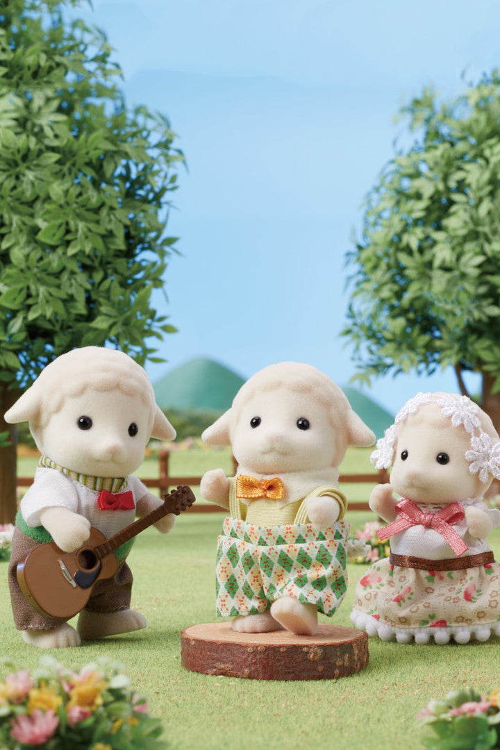 A photo in a pastural scene with 4 sheep. One playing a guitar, one dancing on a stump and 2 red apples and a blue thermos. All the sheep are wearing clothes and they are fleece toys